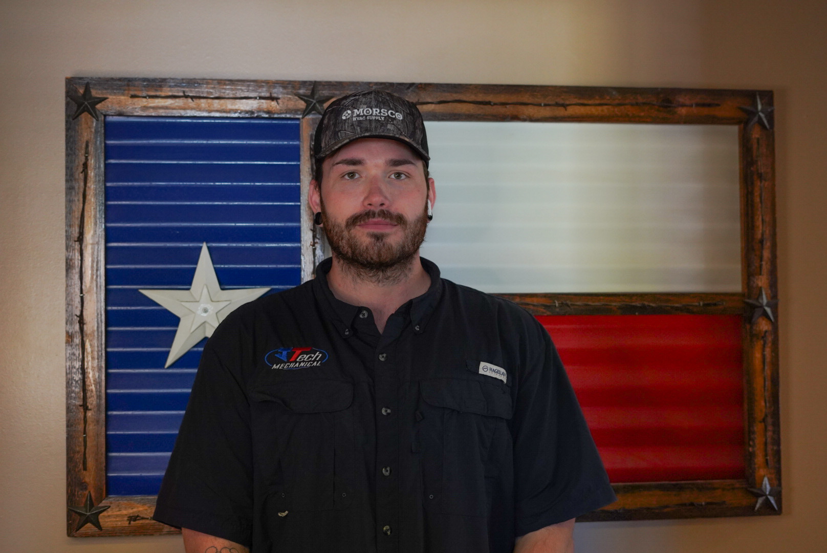Louis – Technician Louis wearing a JTech mechanical shirt standing in front of a metal Texas flag